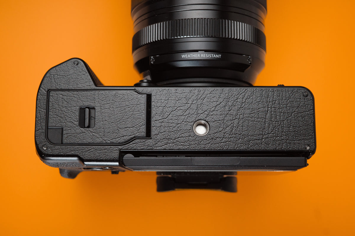 Akara Labs Base Protector. Fujifilm X-T5 black vinyl leatherette-textured camera base. Orange background.