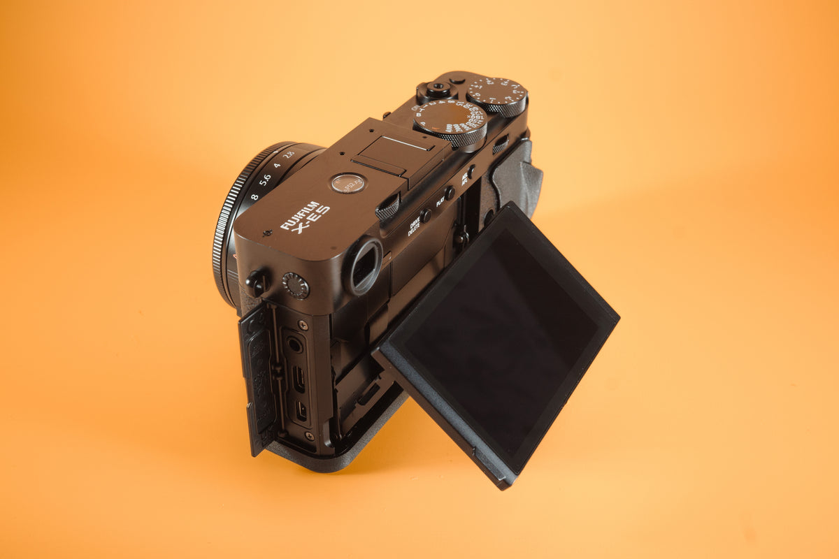 Fujifilm X-E5 in black, rear left view with port door open and screen extended. Fitted with an Akara Labs Slim Skin grip case in black leatherette-textured TPU