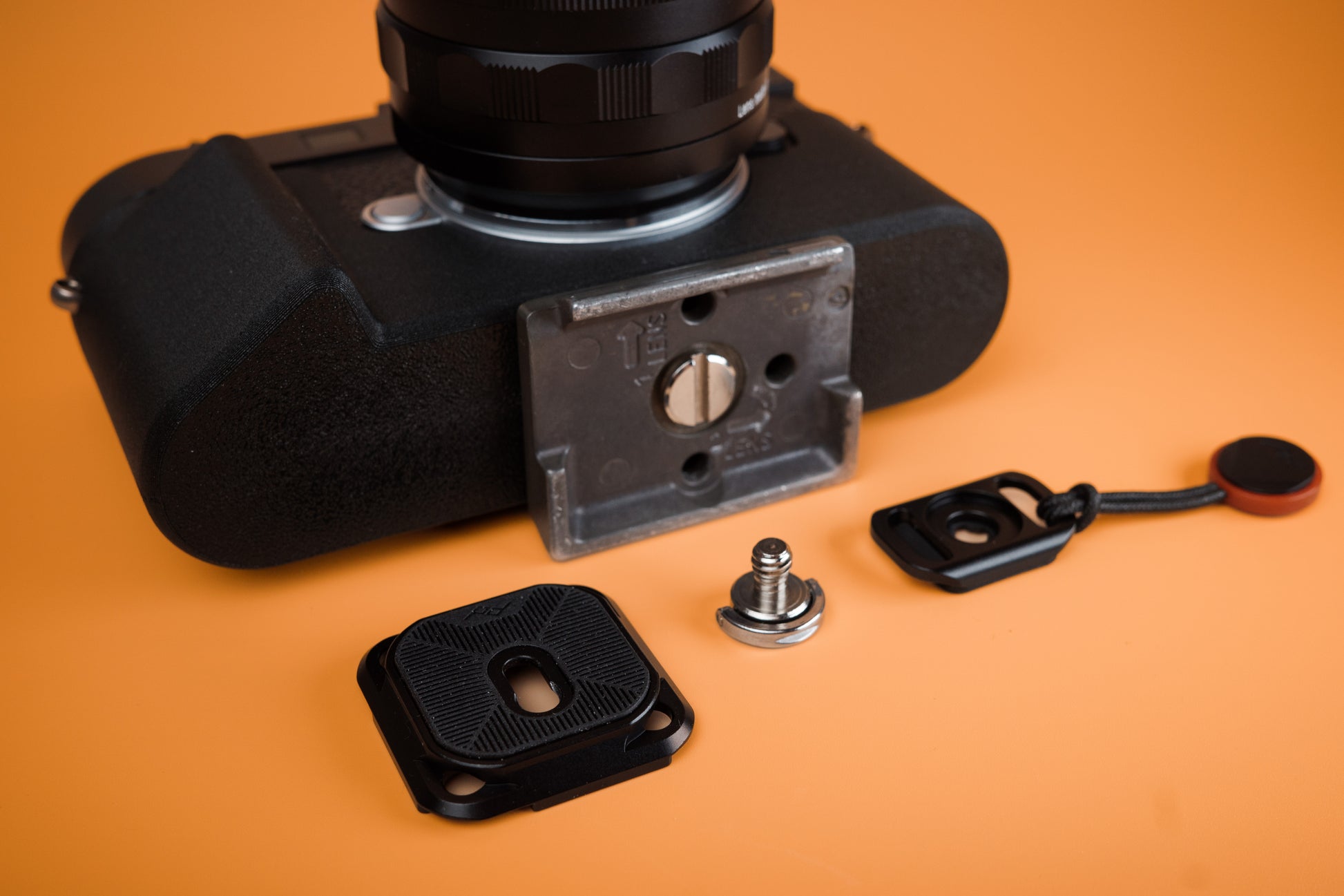 Leica M11 TPU Slim Skin case. Bottom view with tripod attachment. Peak Design mounting hardware laid on orange background.
