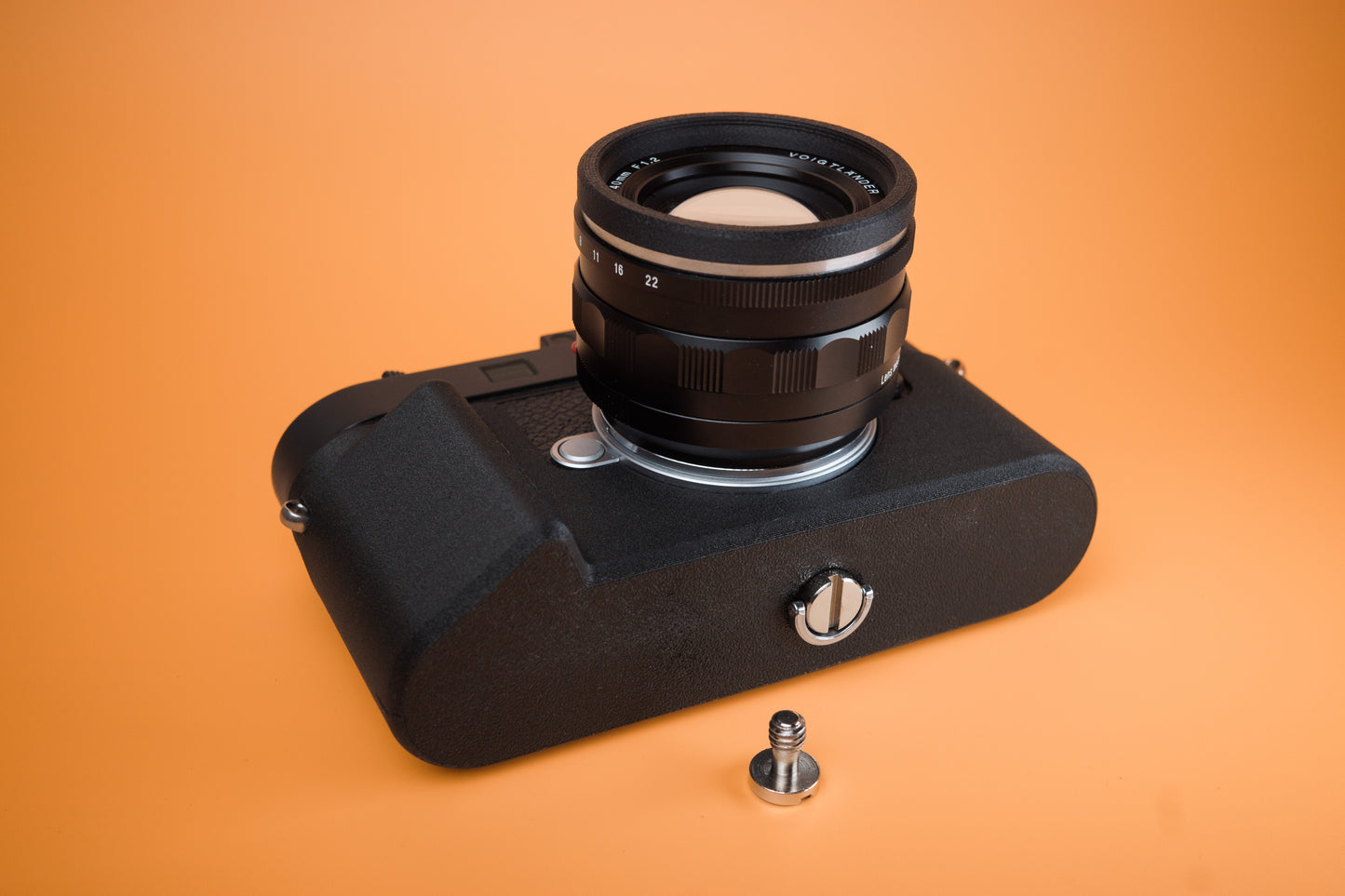 Leica M11 Slim Skin Tripod version case with two mounting screws. Bottom view with camera laying on rear. Orange background.