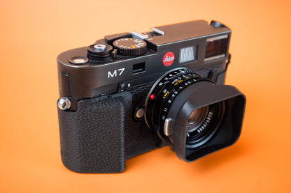 A black Leica M7 rangefinder camera with Mandler 35mm lens and a front lens hood, in a Akara Labs Slim Skin Grip case. photographed on a solid orange background. 