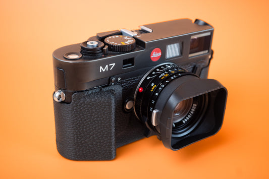 A black Leica M7 rangefinder camera with Mandler 35mm lens and a front lens hood, in a Akara Labs Slim Skin Grip case. photographed on a solid orange background. 