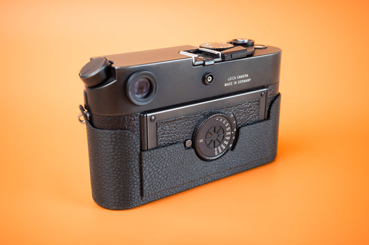 A vintage Leica M7 rangefinder camera with a black textured Slim Skin case and black metal body, photographed from a three-quarters rear left angle against a solid orange background.