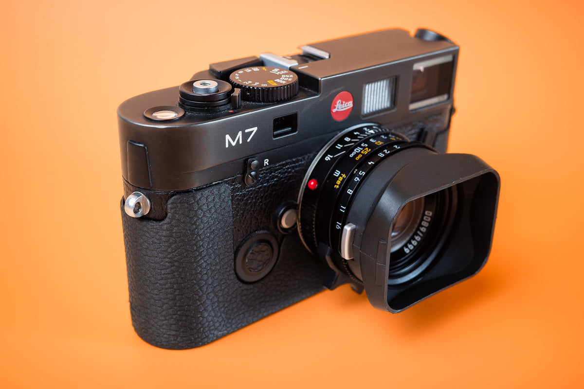 Leica M7 black leatherette-textured style Slim Skin standard case. Front right view, photographed against an orange background.